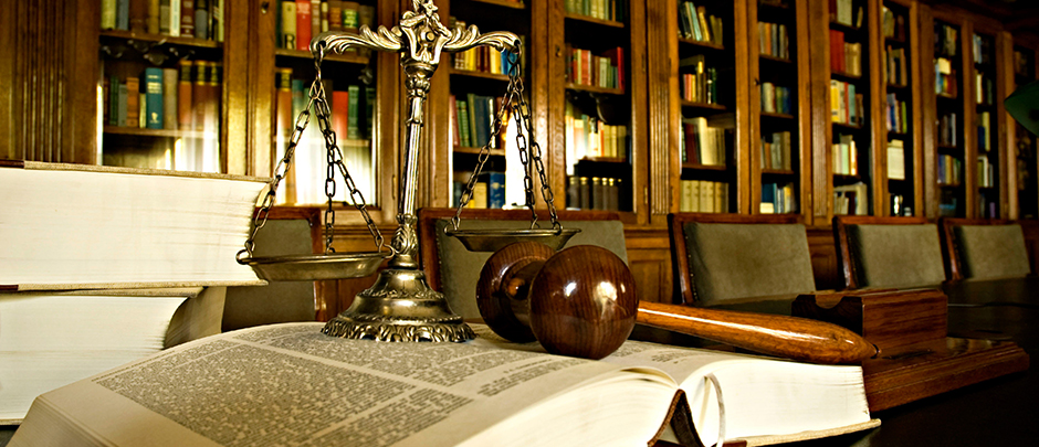  An image of a gavel and scales of justice on a desk in a library with a law firm meeting in the background.